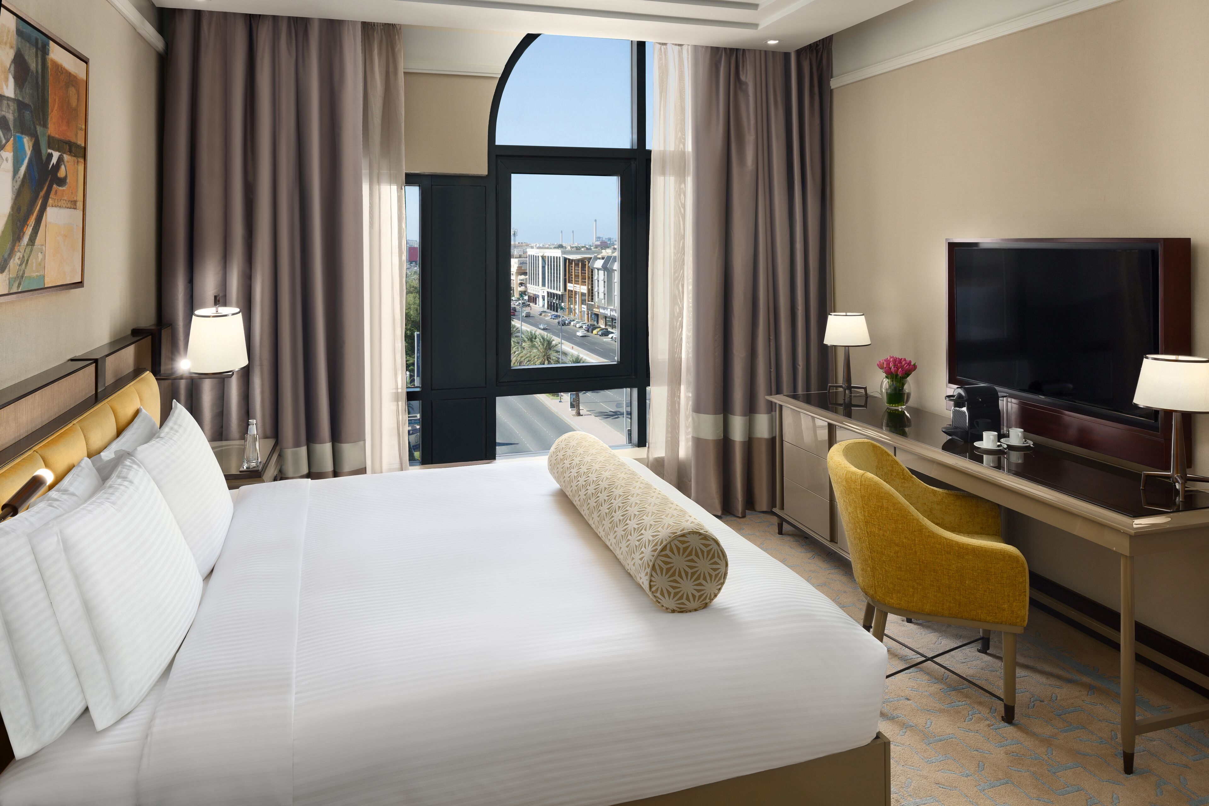 Quarto de hotel com uma cama grande, uma escrivaninha com televisão e vista para a cidade através de uma janela ampla.