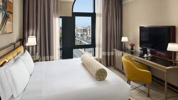 Quarto de hotel com uma cama grande, uma escrivaninha com televisão e vista para a cidade através de uma janela ampla.