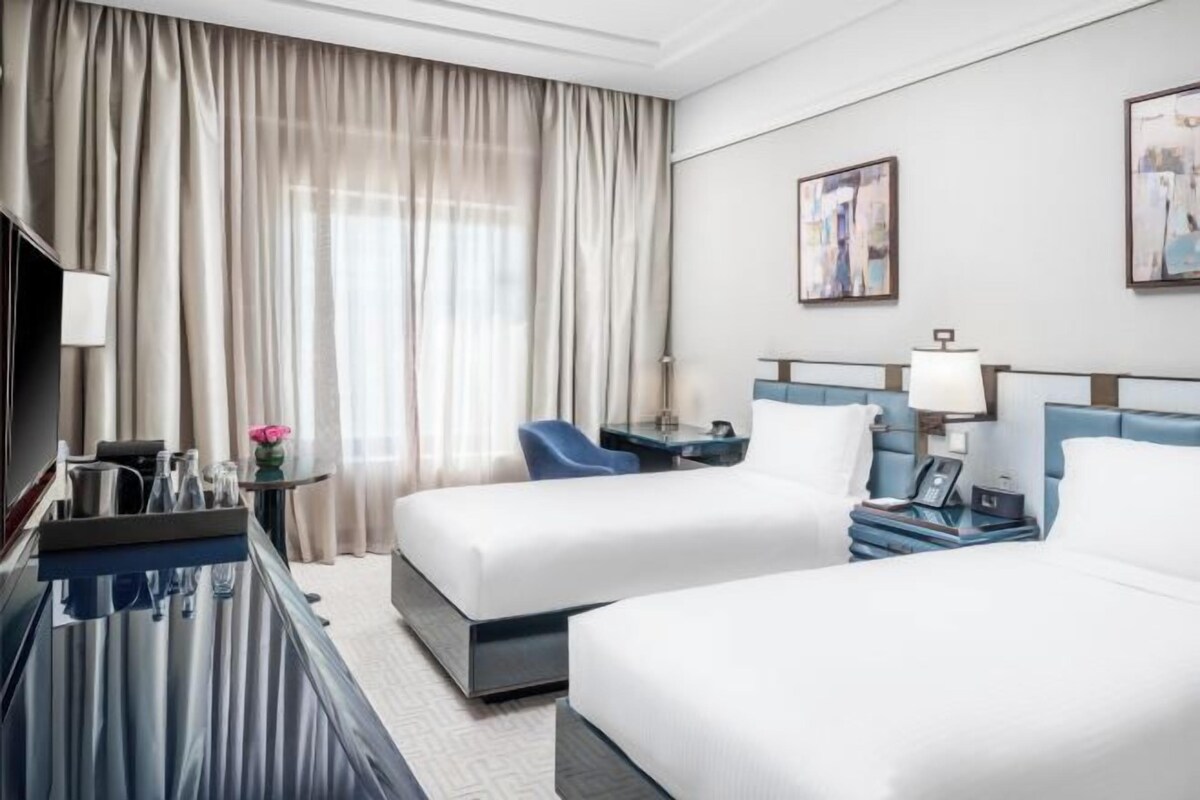 A hotel room with two beds, a desk, a chair, a TV, and a window with curtains.