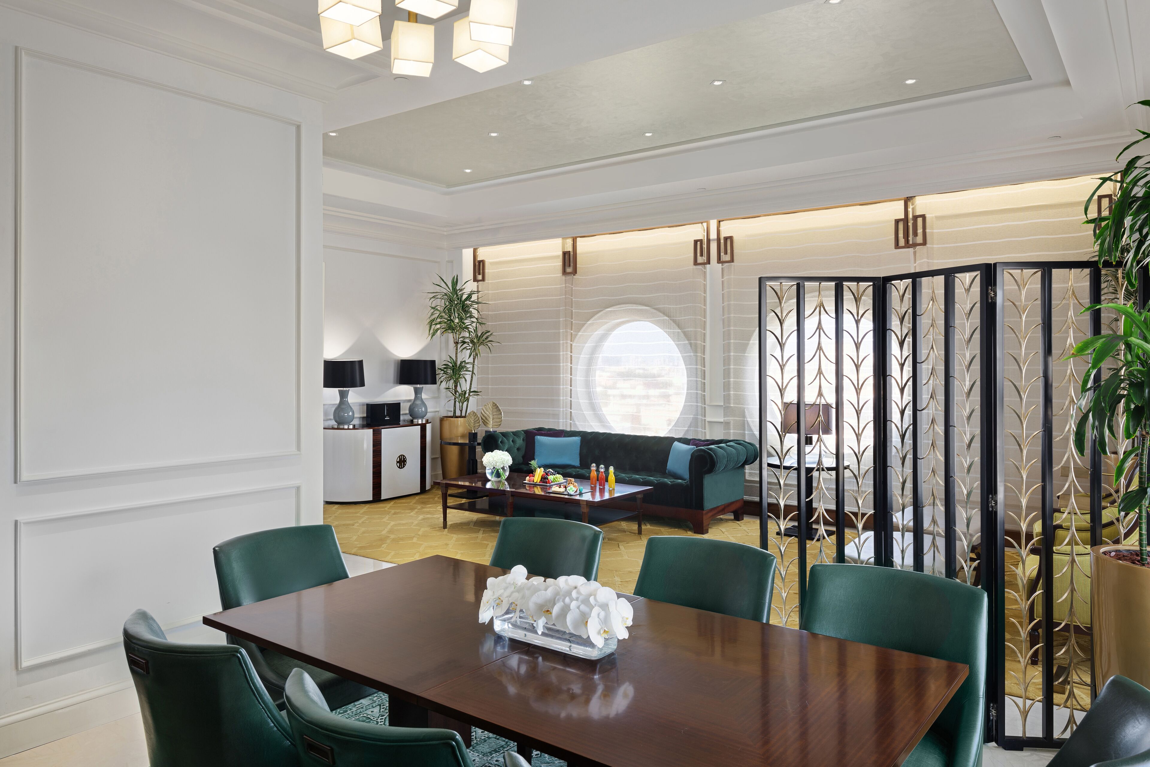 A modern dining area with a wooden table and green upholstered chairs, a sofa, and a decorative partition with a circular window.