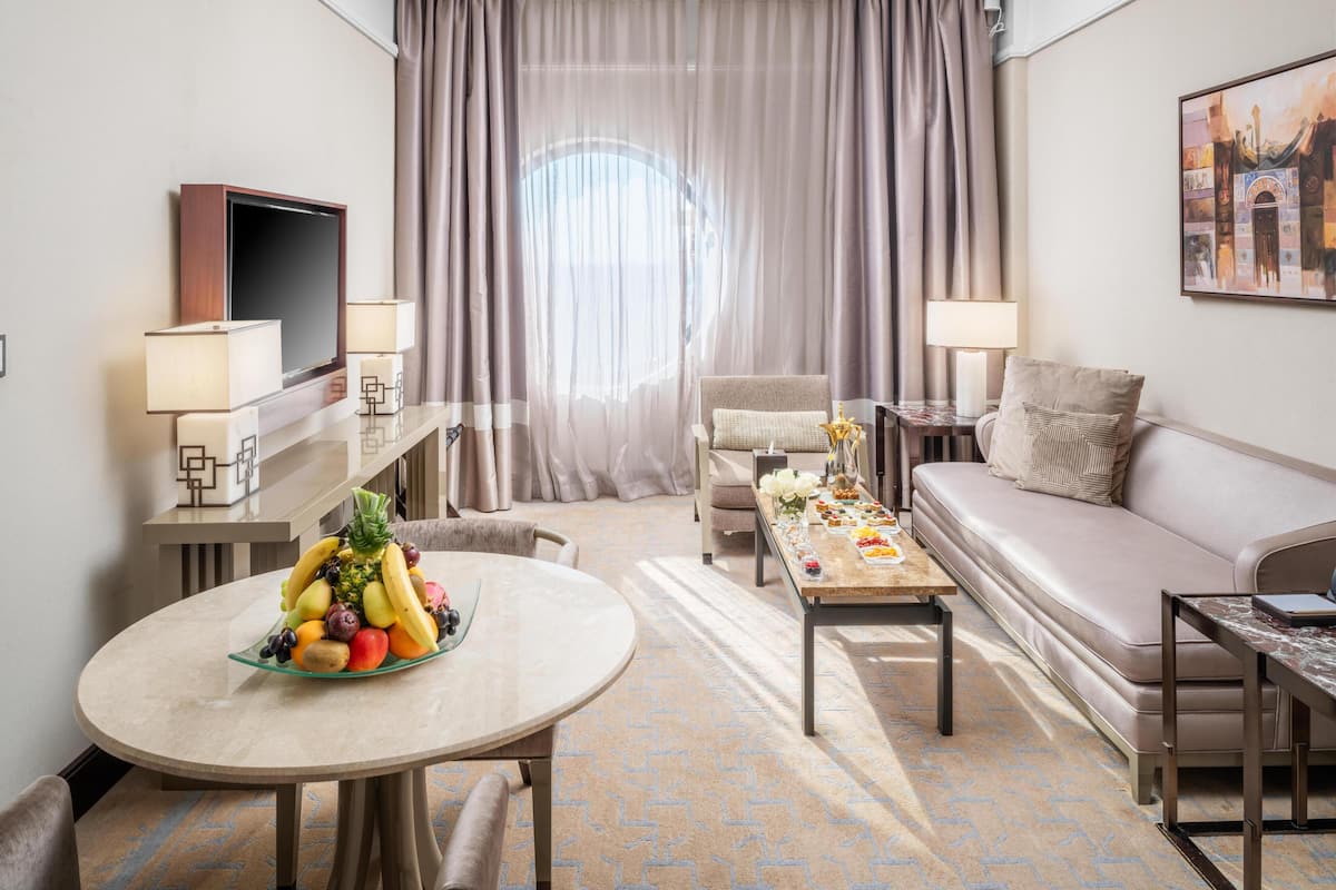 Een hotelkamer met een ronde eettafel, een bank, een televisie en een groot raam met gordijnen.