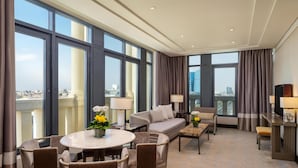 Habitación de hotel con un ventanal grande, un sofá, una mesa de comedor con sillas, un televisor y una pequeña mesita de noche con lámpara.