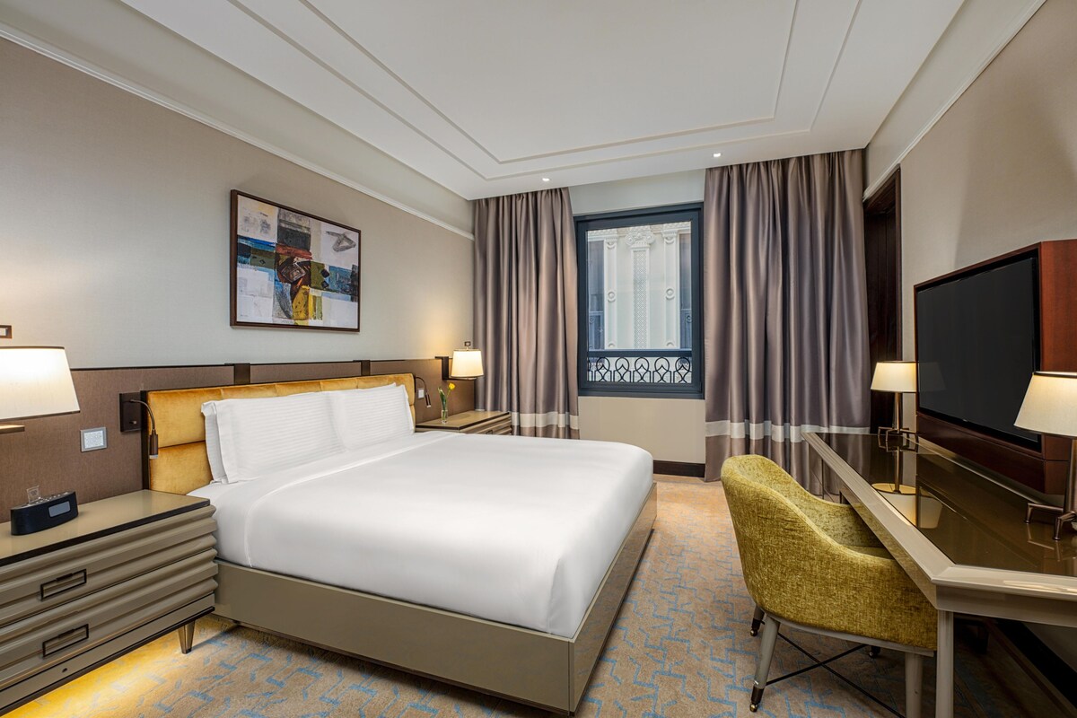 Una habitación de hotel con cama, escritorio, televisión y ventana con cortinas.