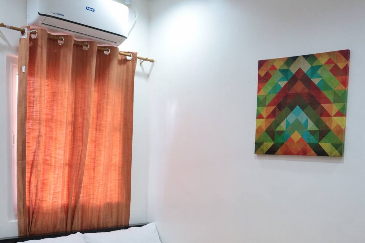 A single bed with a beige mattress and white pillows, an air conditioning unit above, and a colorful geometric painting on the wall.
