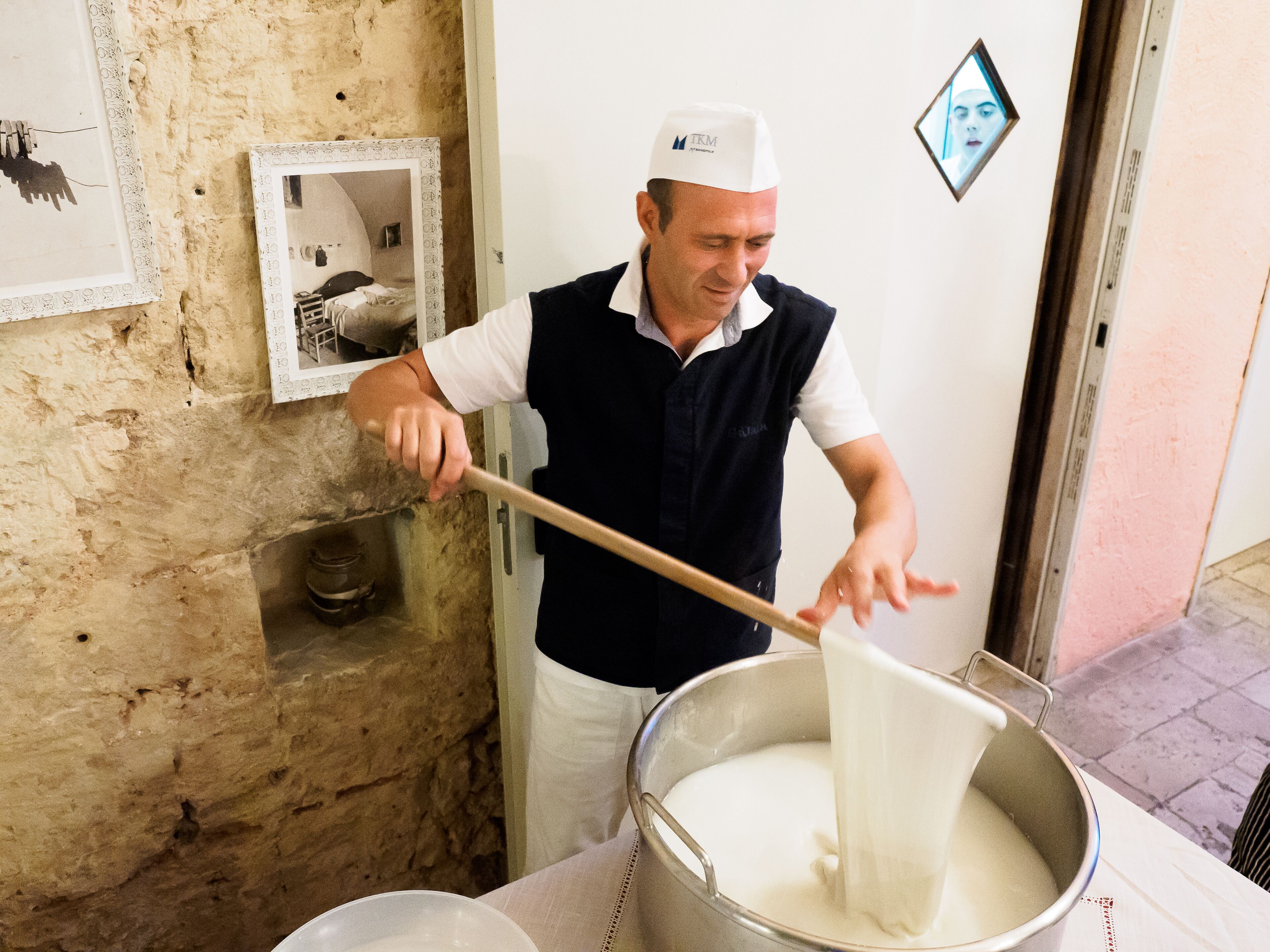 A person stirring a large pot of white liquid, likely milk or cream, in a kitchen setting with a rustic stone wall and a framed picture on the wall.