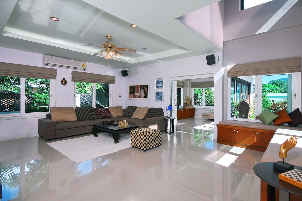 A spacious living room with a large sofa, a coffee table, and a checkered ottoman. There is a ceiling fan, a window with a view of greenery, and a statue on a table.