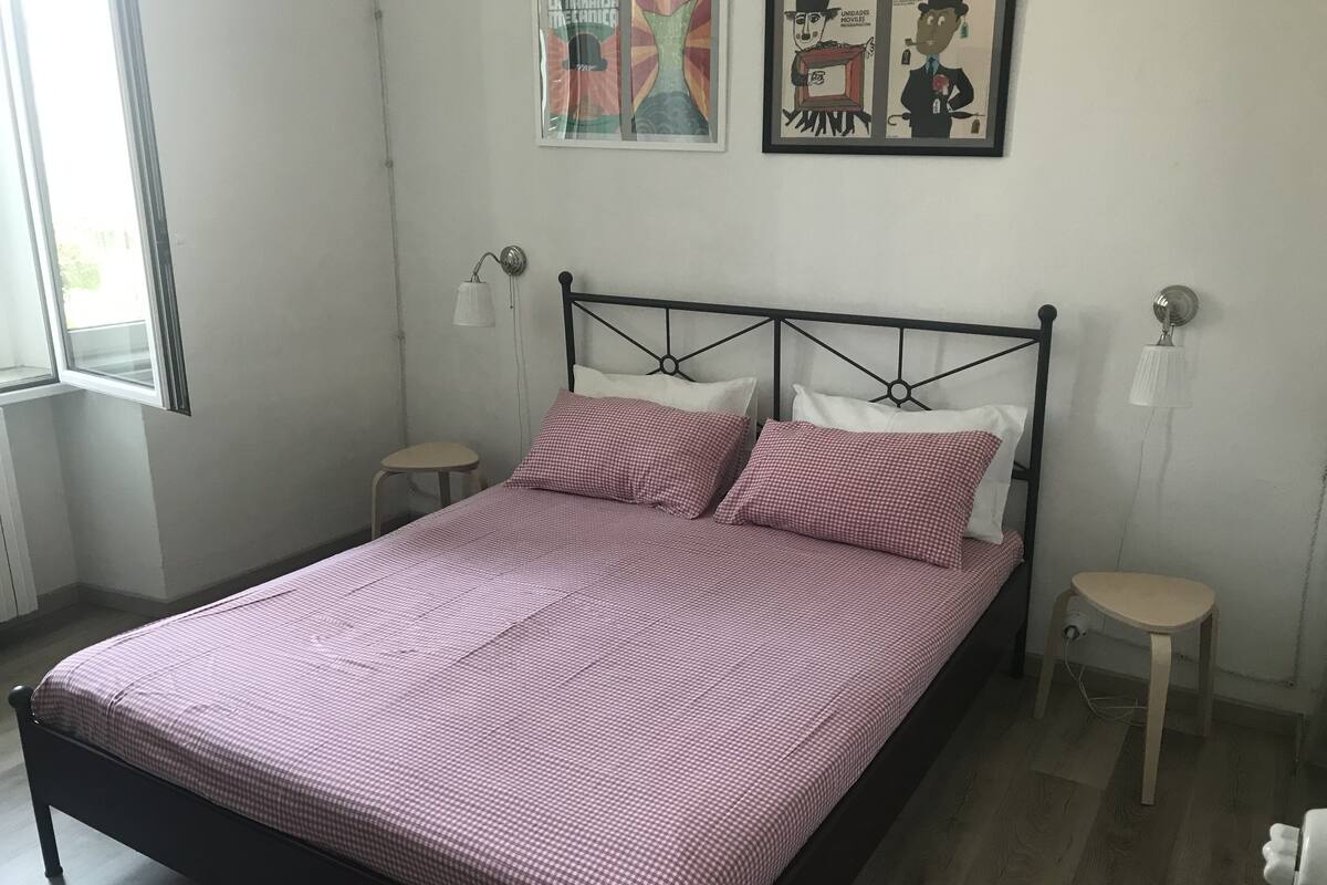 A neatly made bed with a checkered bedspread, two bedside tables, a wall-mounted lamp, and two framed artworks above the bed.