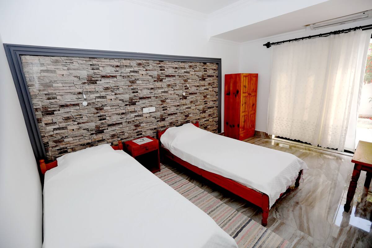 Habitación de hotel con dos camas individuales, cabecero de madera, armario rojo y una ventana grande con cortinas.