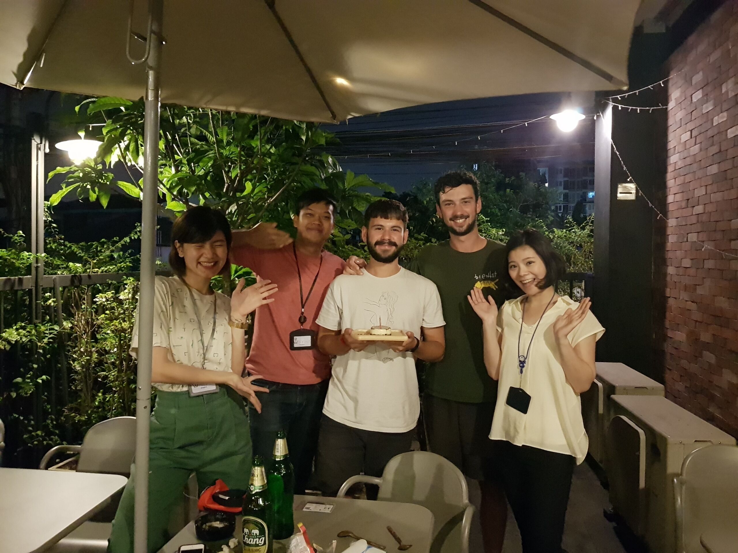 Un grupo de personas de pie bajo un paraguas, con una mesa y sillas al frente.