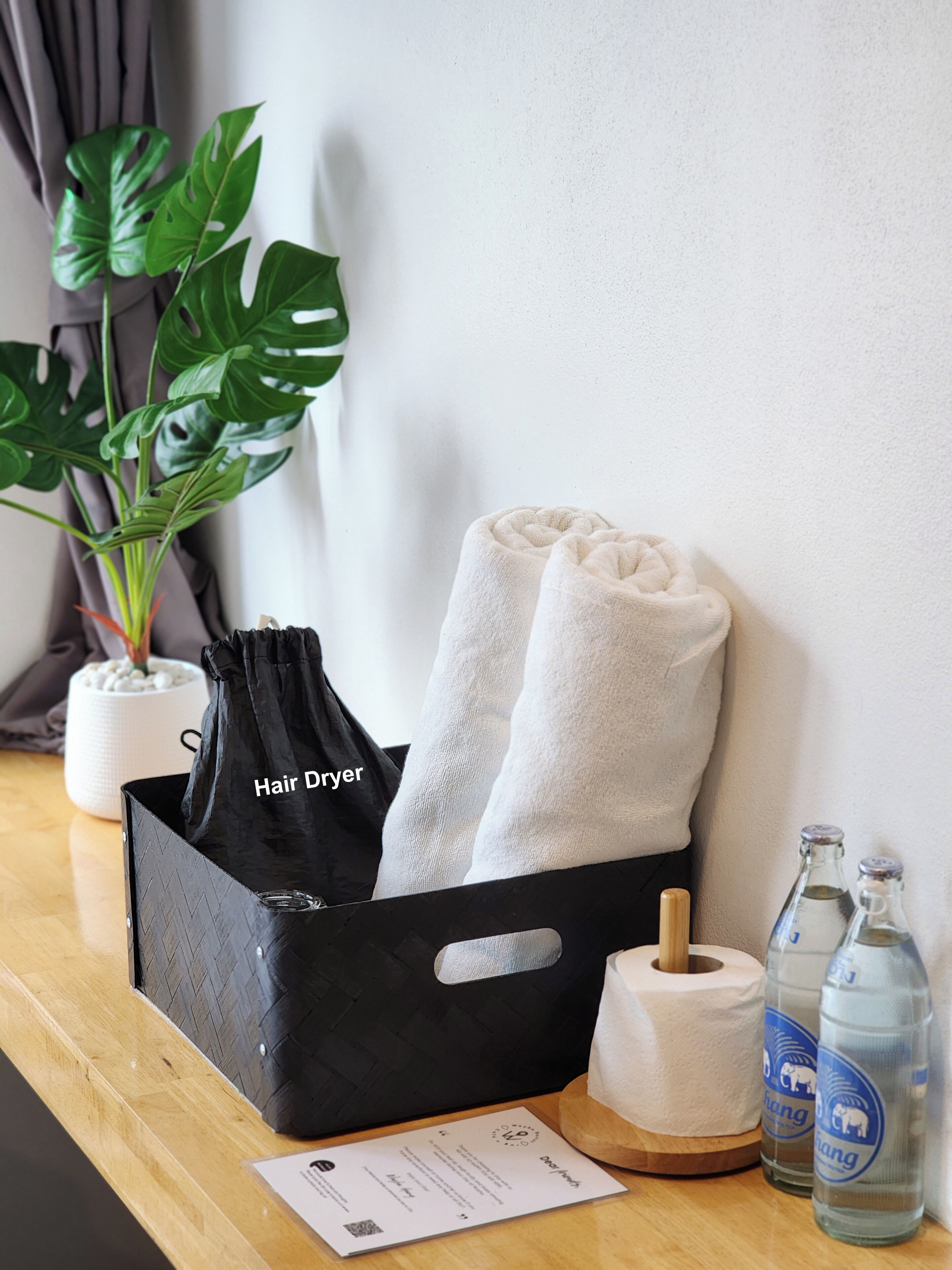 Un sac noir pour sèche-cheveux, deux serviettes blanches, un rouleau de papier toilette et deux bouteilles d’eau sur une table en bois.