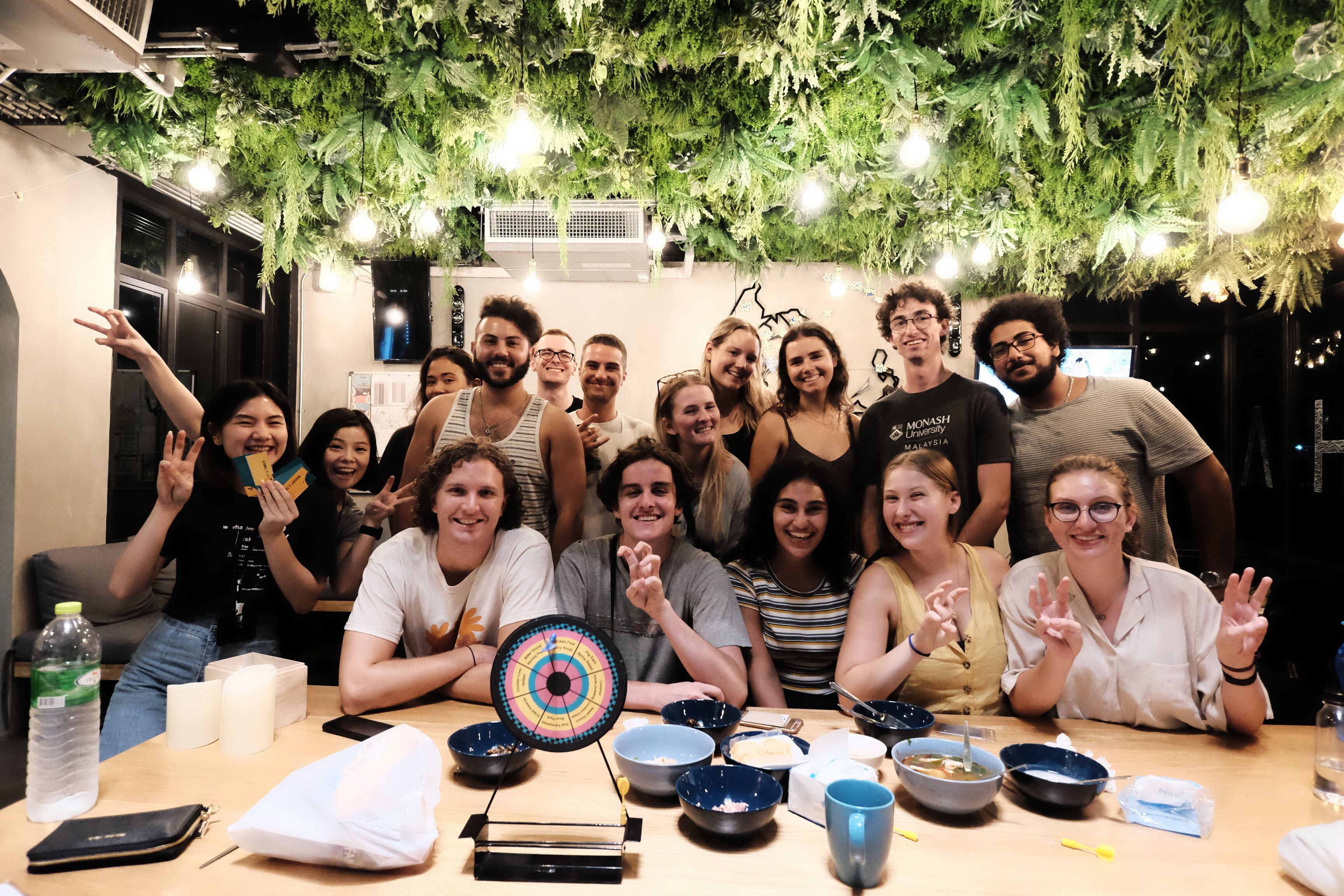 A group of people seated around a table with bowls and a spinning wheel, in a room with hanging greenery and pendant lights.