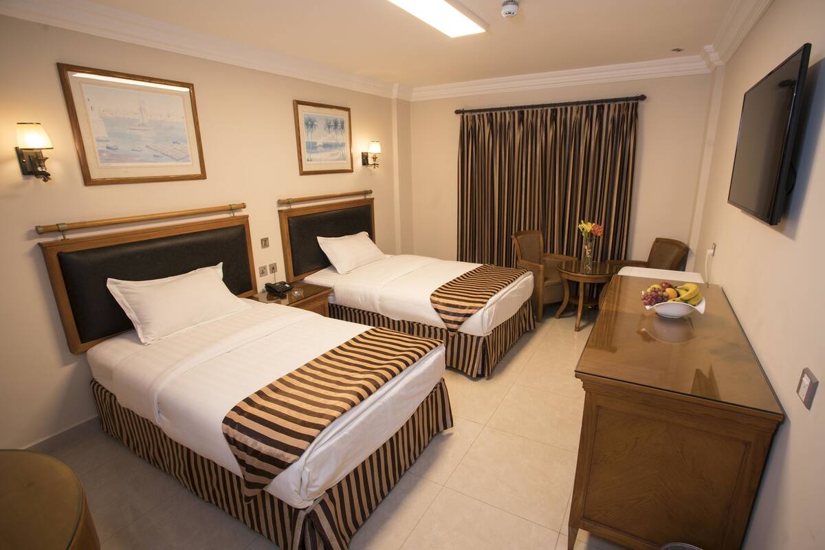 A hotel room with two beds, a desk, a chair, a TV, and a dining table with fruit.