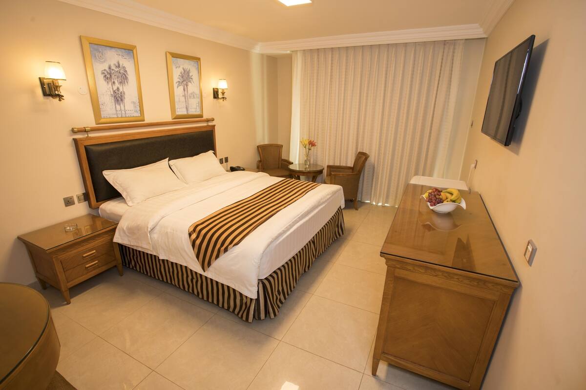 A hotel room with a large bed, a wooden nightstand, a desk with a bowl of fruit, and a television on the wall.