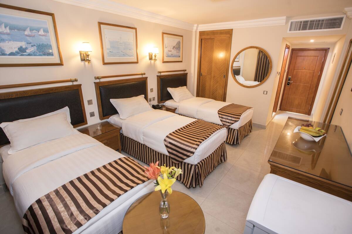 A hotel room with three beds, a round mirror, a small table with a vase of flowers, and a wooden door.