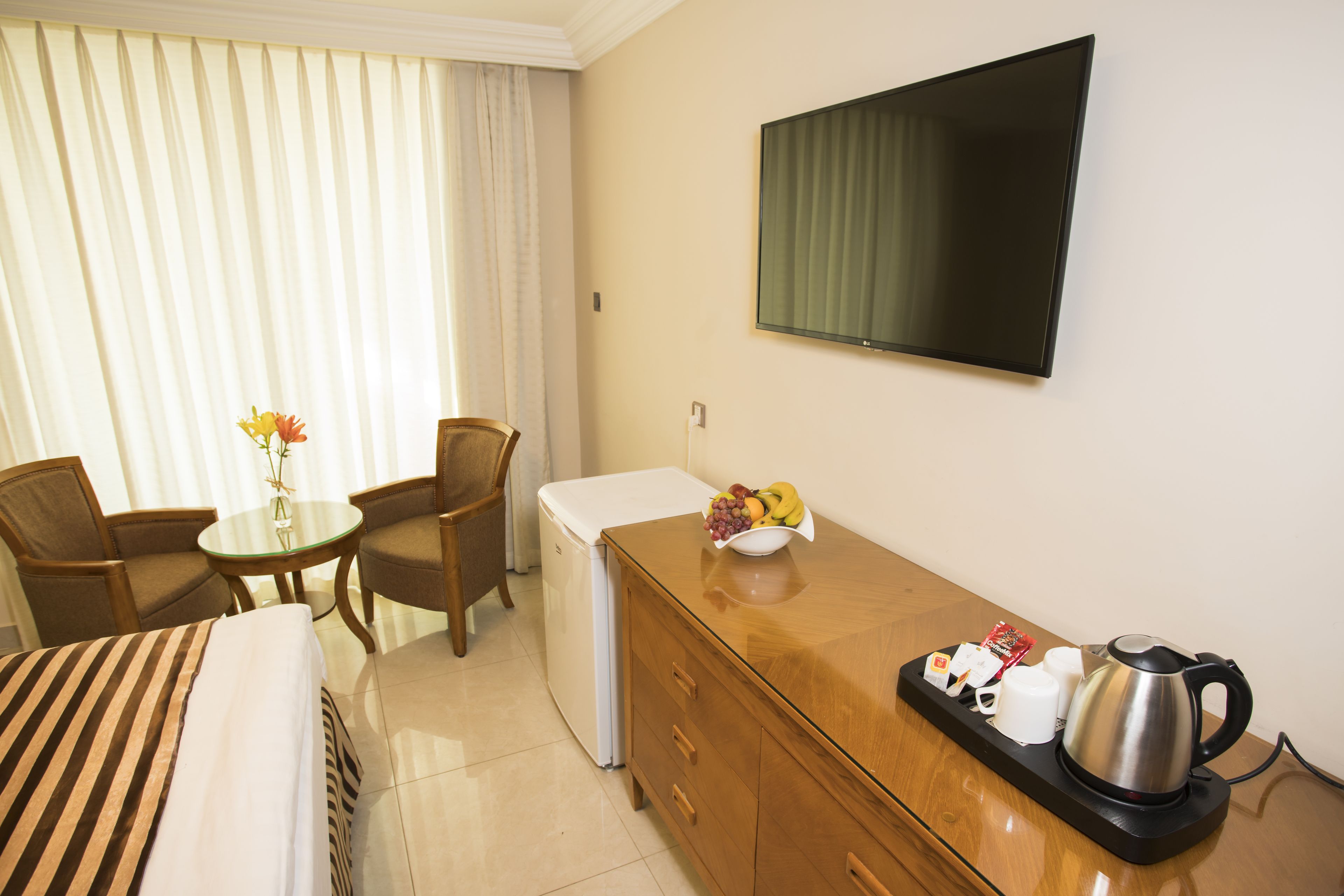 Ein Hotelzimmer mit Bett, kleinem Tisch mit Stühlen, Kühlschrank, Holzkommode mit Frühstücksprodukten und flachbildschirmfernseher an der Wand.