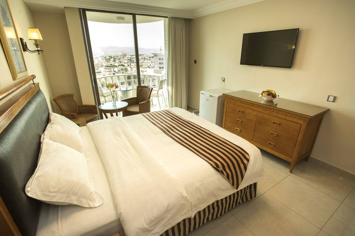 A hotel room with a bed, a wooden dresser, a television, and a balcony with a view of the city.