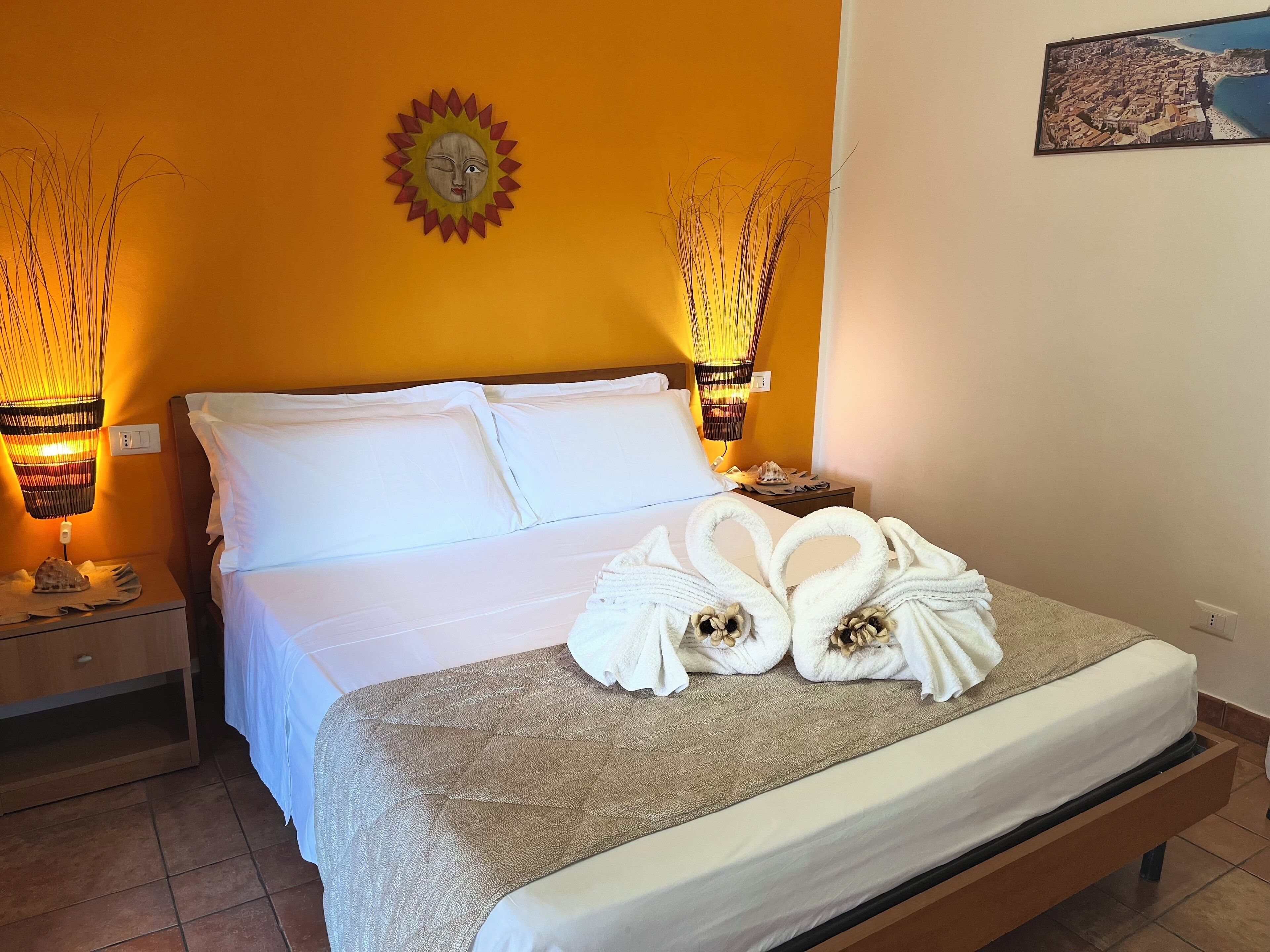 A hotel room with a bed, two bedside lamps, a wall-mounted sun decoration, and a framed picture.