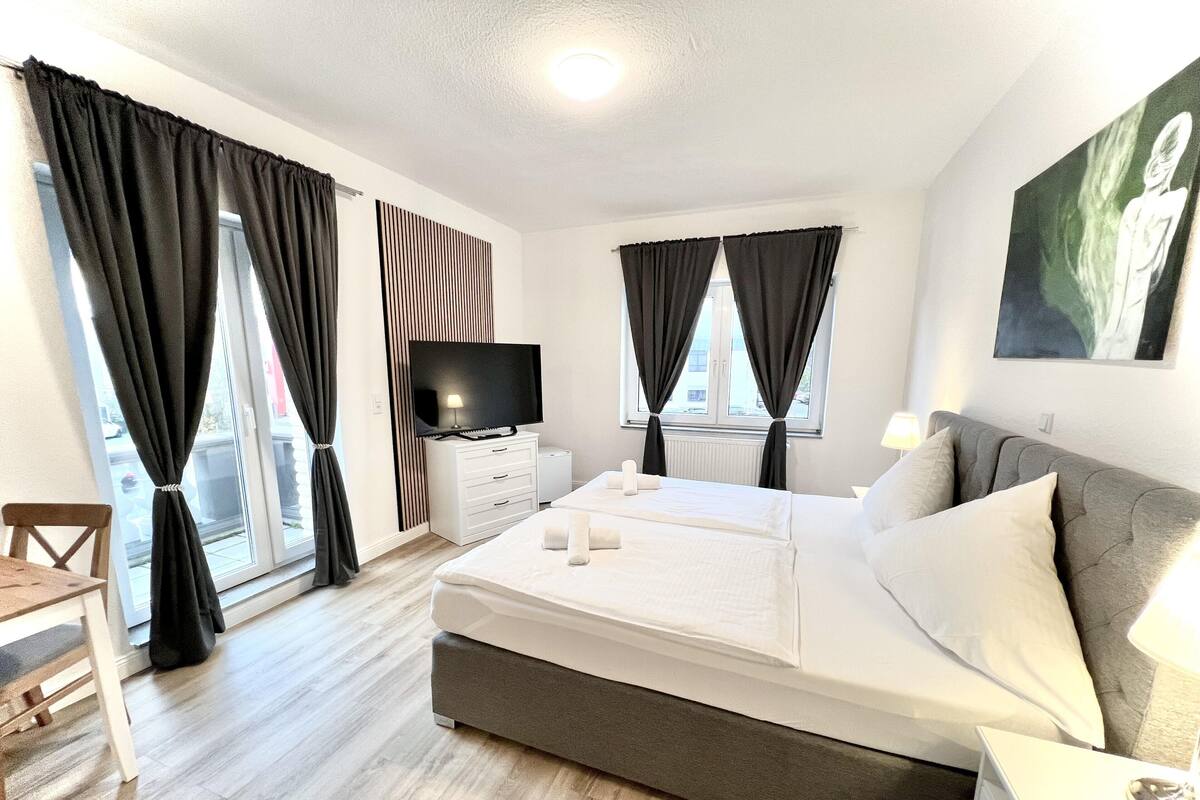A modern hotel room with a bed, bedside tables, a TV, and large windows with curtains.
