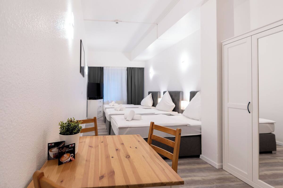 A modern hotel room with a wooden dining table, a potted plant, and a bed with white linens.