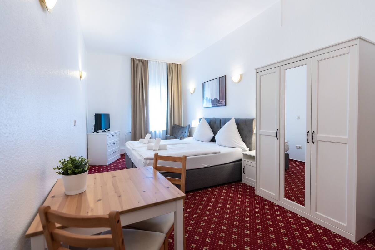 A hotel room with a bed, bedside table, chair, wardrobe, and a desk with a plant.