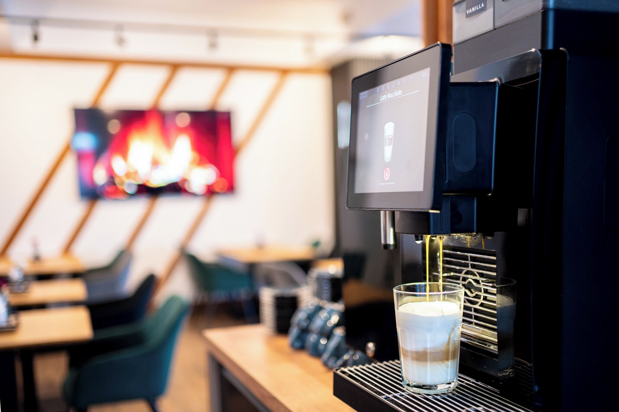 A coffee machine dispensing coffee into a glass, with a digital display and a television in the background.