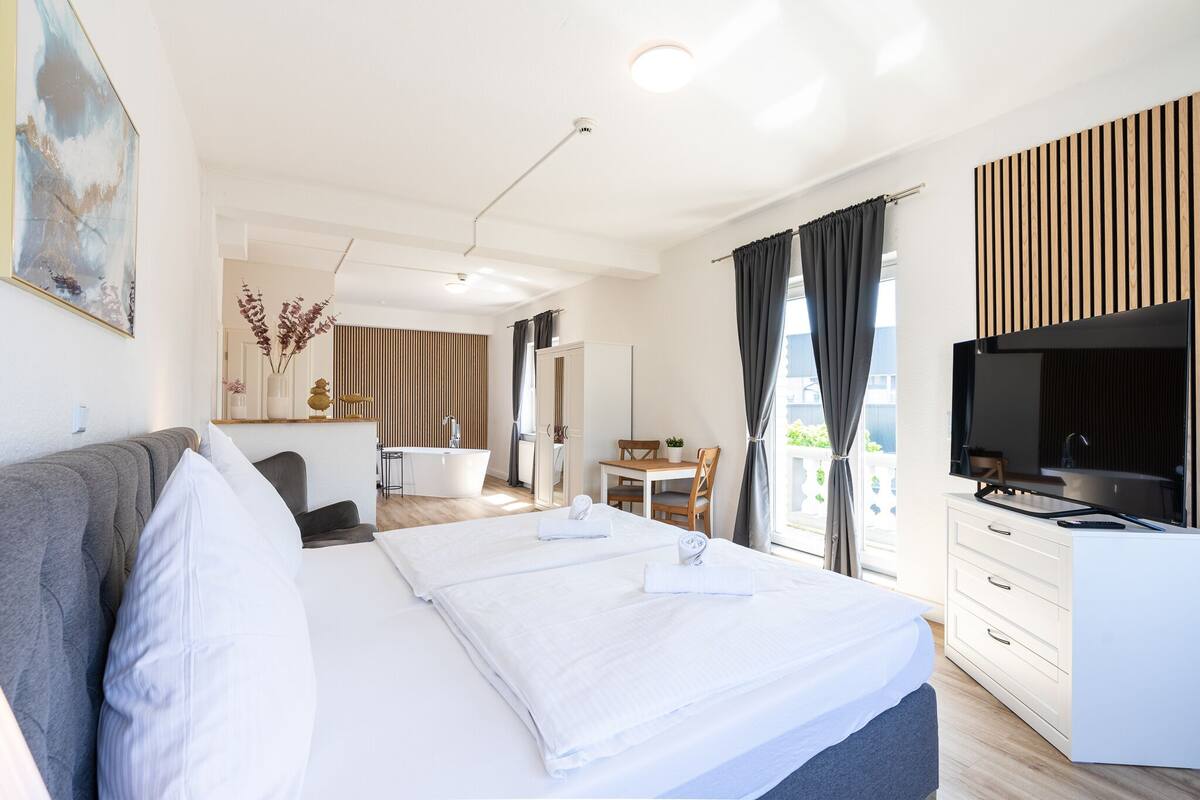 A modern hotel room with a large bed, a desk, a TV, and a bathroom visible in the background.