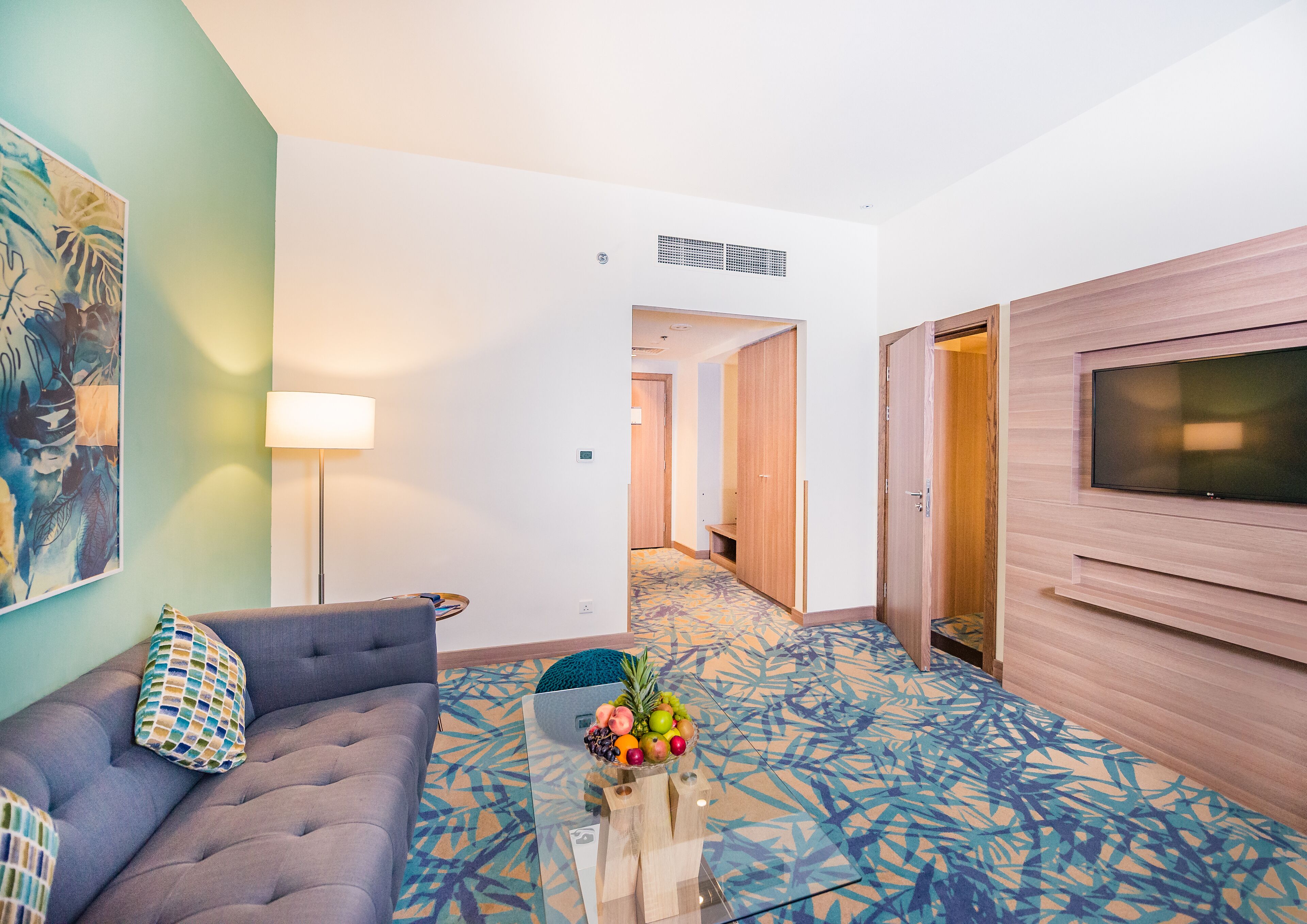 A modern hotel room with a sofa, a coffee table with fruit, a TV, and a patterned carpet.