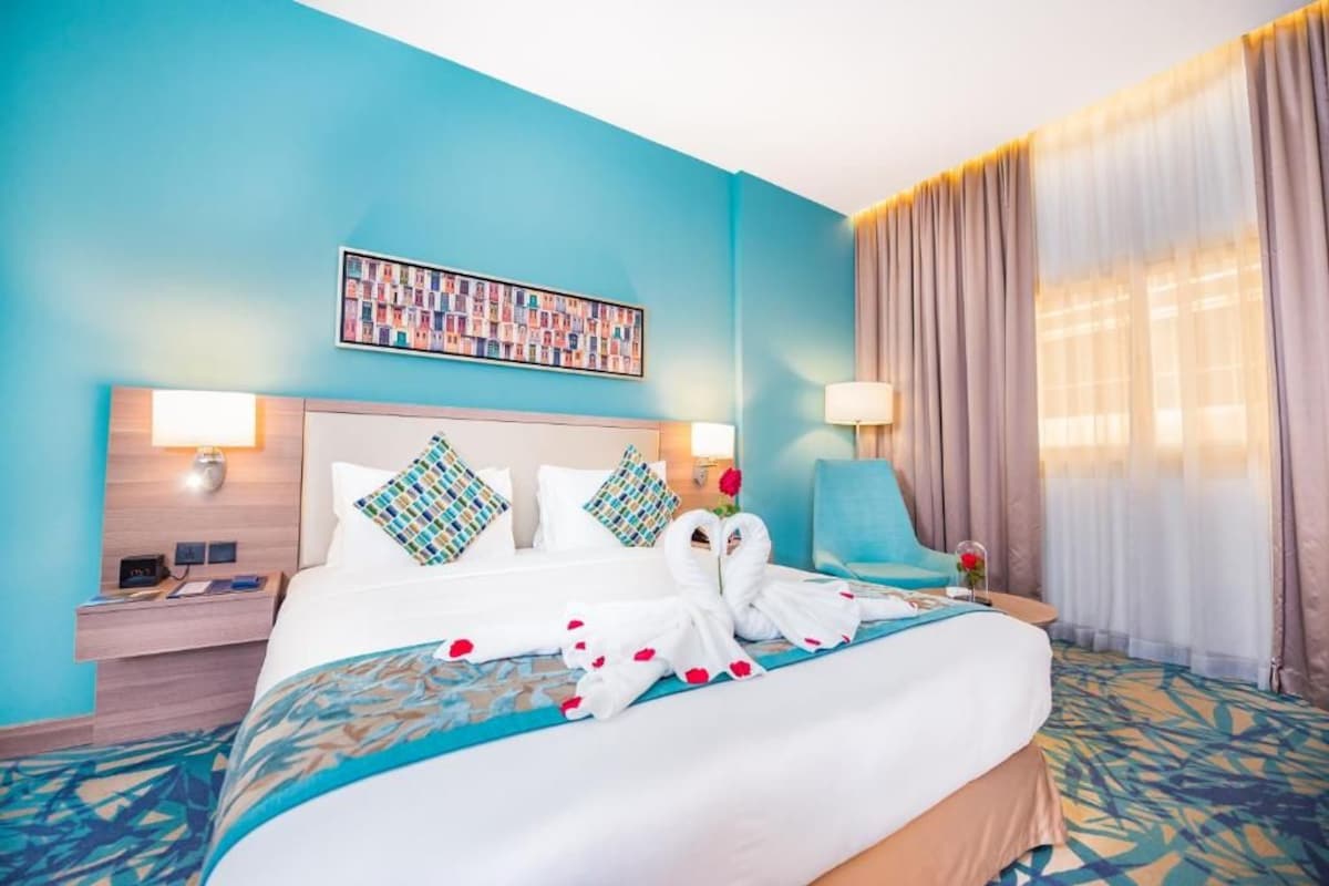 Habitación de hotel con una cama grande, dos almohadas, una mesita de noche, una lámpara, una silla y una ventana con cortinas.