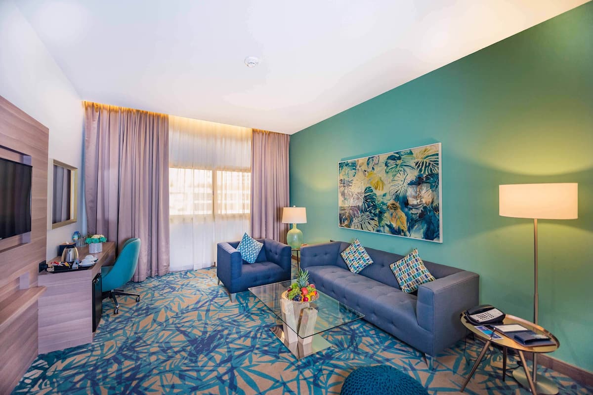 Een moderne hotelkamer met een bank, een bijzettafeltje met fruit, een bureau en een groot raam met gordijnen.