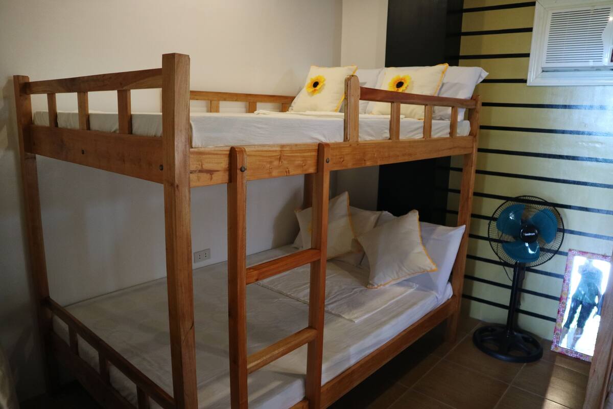 A bunk bed with a wooden frame, white bedding, and sunflowers on pillows. A standing fan is next to the bed.