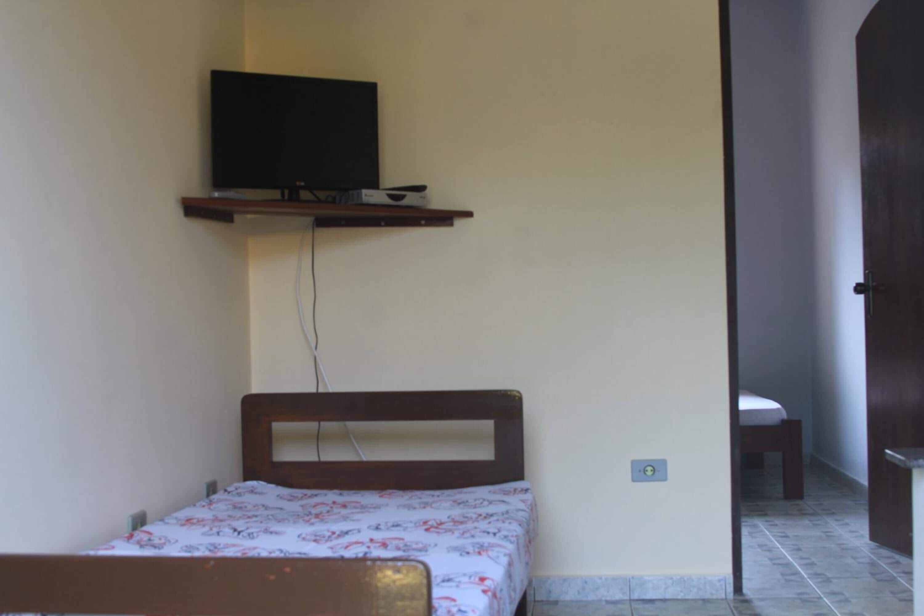 Een kleine, eenvoudige kamer met een bed, een aan de muur bevestigde televisie en een deur naar een andere kamer.
