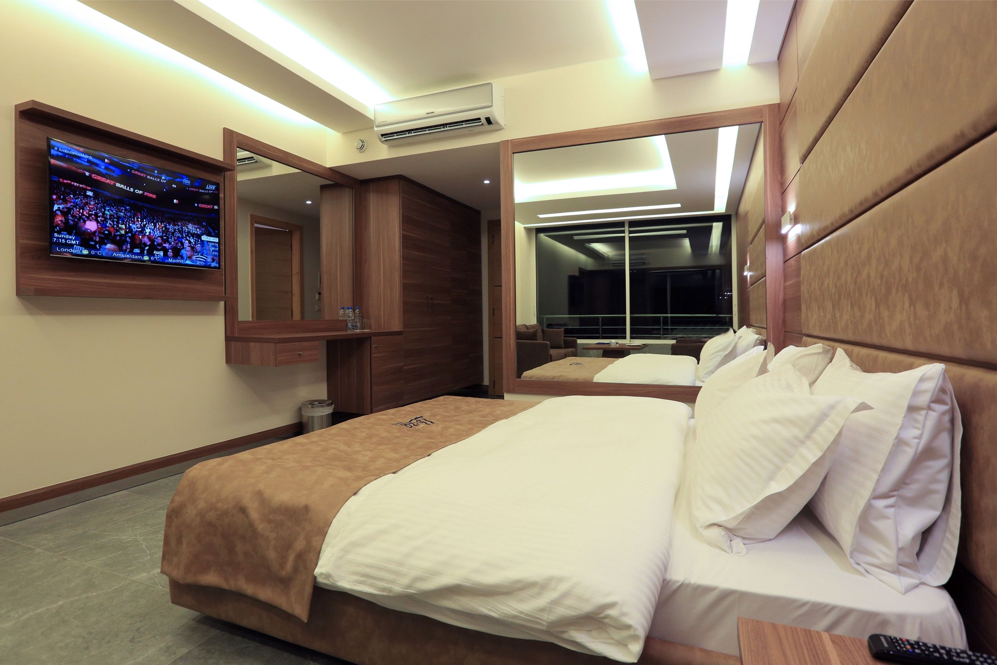 A hotel room with a large bed, a TV mounted on the wall, a mirror, and an air conditioning unit.