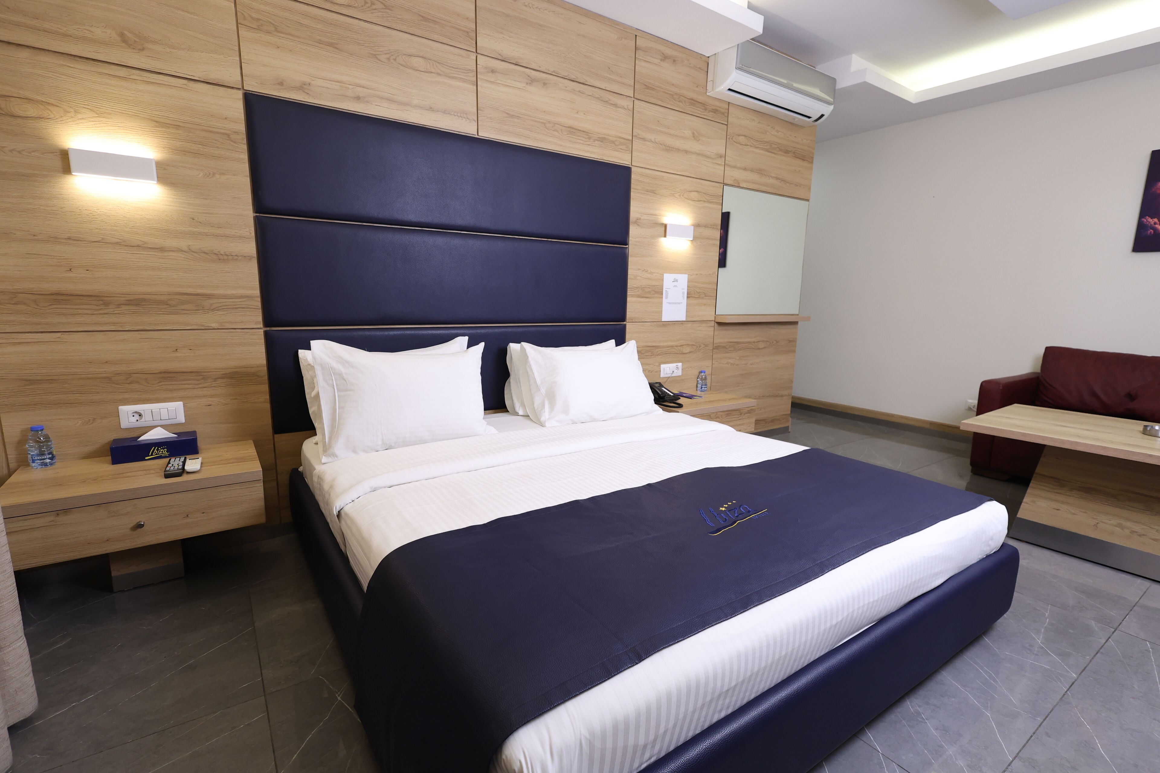 A modern hotel room with a large bed, wooden panel walls, a nightstand with a water bottle, and a desk with a chair.