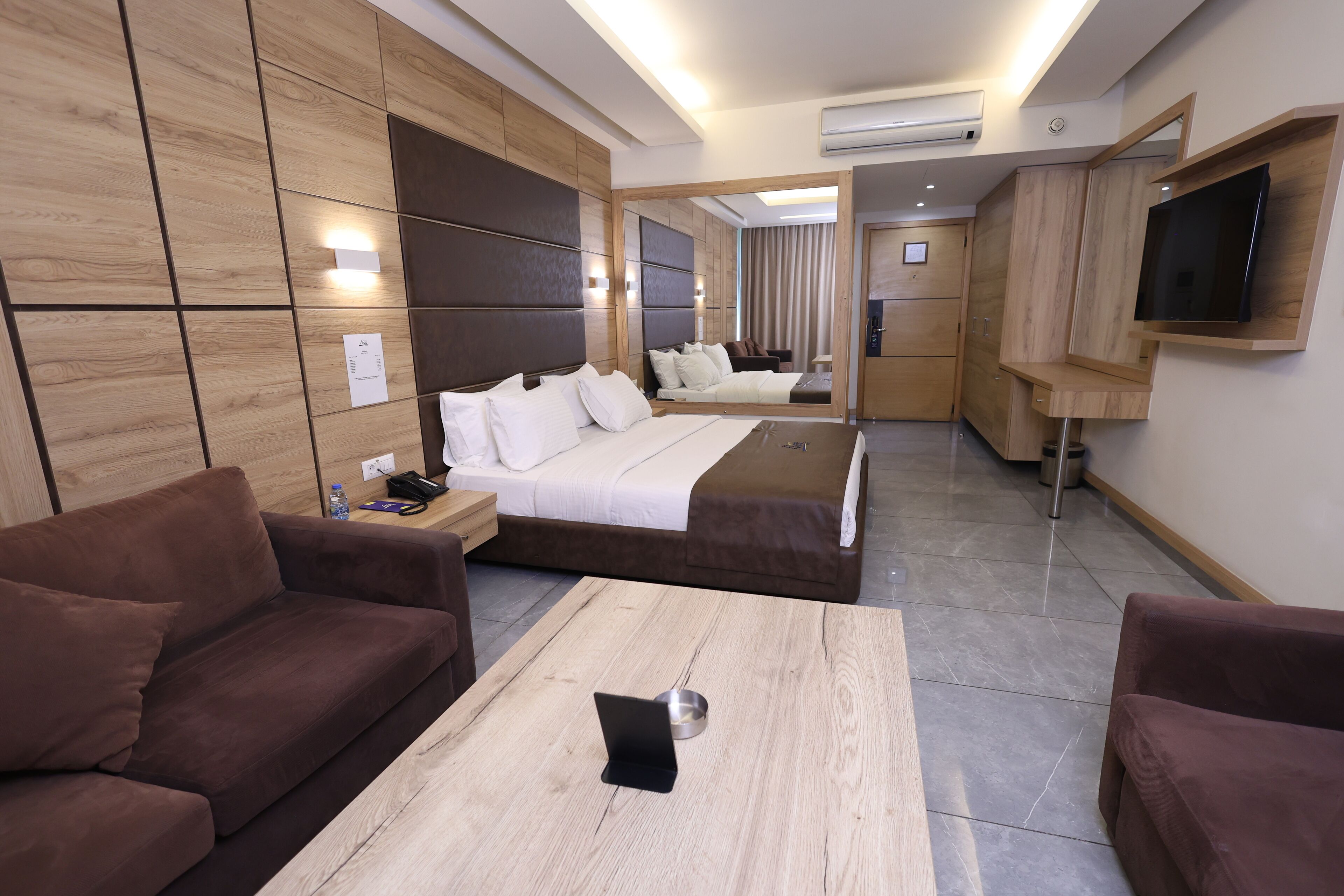 Une chambre d’hôtel moderne avec un grand lit, deux canapés marron, une table basse en bois et un téléviseur fixé au mur.