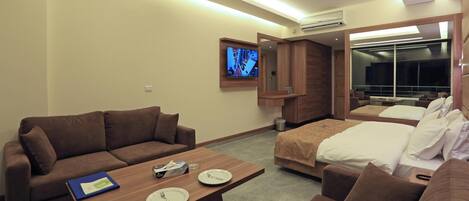 A hotel room with a brown sofa, a wooden coffee table with plates and a book, a bed with white bedding, and a TV mounted on the wall.