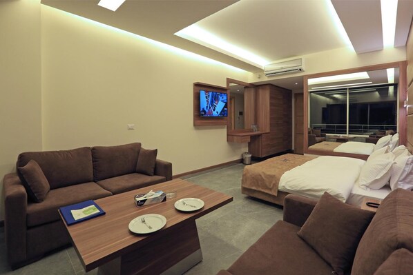 Quarto de hotel com um sofá castanho, uma mesa de centro de madeira com pratos e um livro, uma cama com roupa de cama branca e uma televisão fixada na parede.