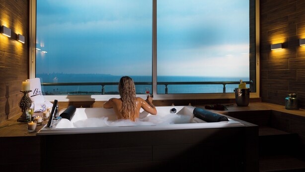 A woman relaxing in a bathtub with a view of the sea through large windows.