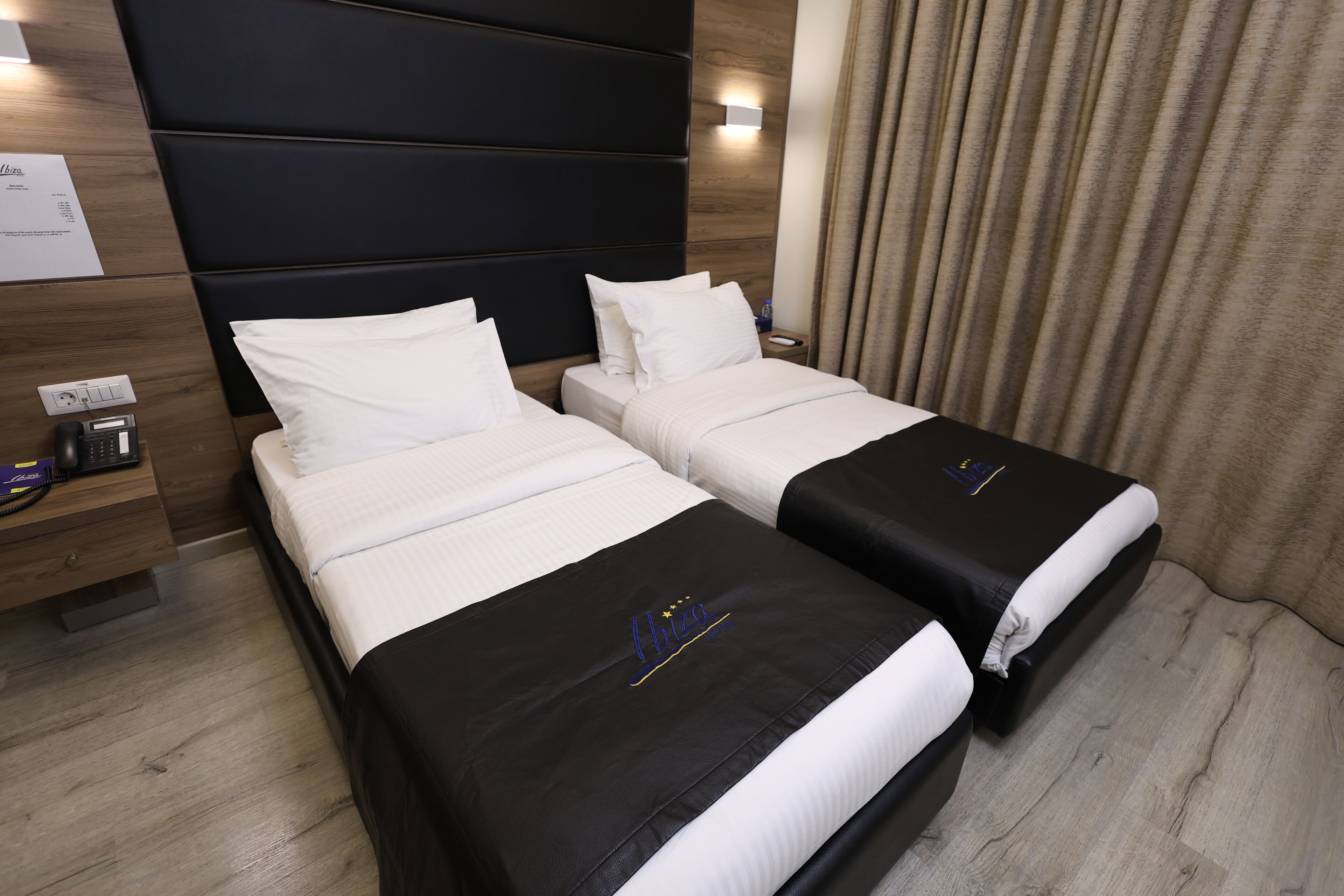 Una habitación de hotel con dos camas, un cabecero de madera, una mesita de noche con una lámpara y una cortina.