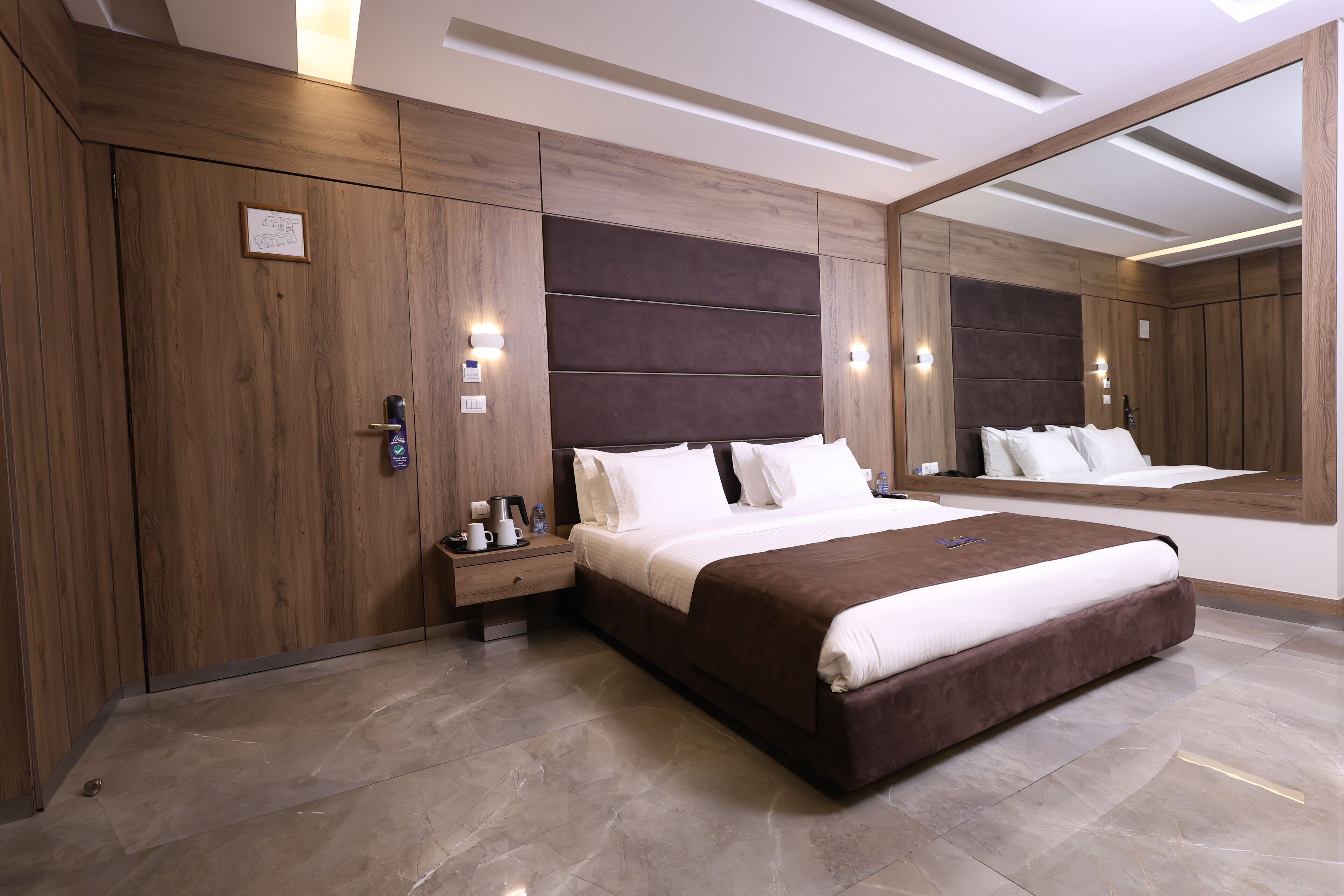 A modern bedroom with a large bed, wooden paneling, a nightstand with a lamp, and a large mirror.