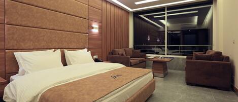 A modern hotel room with a large bed, two armchairs, a small table, and a view of the city through a large window.