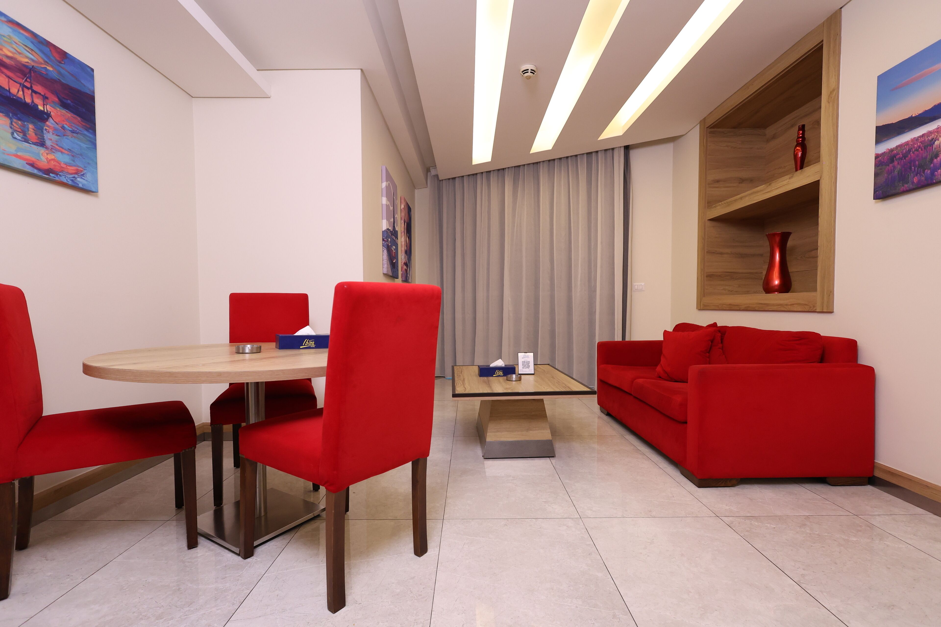 A modern living room with a red sofa, a red dining table with chairs, and a wooden shelf with decorative items.