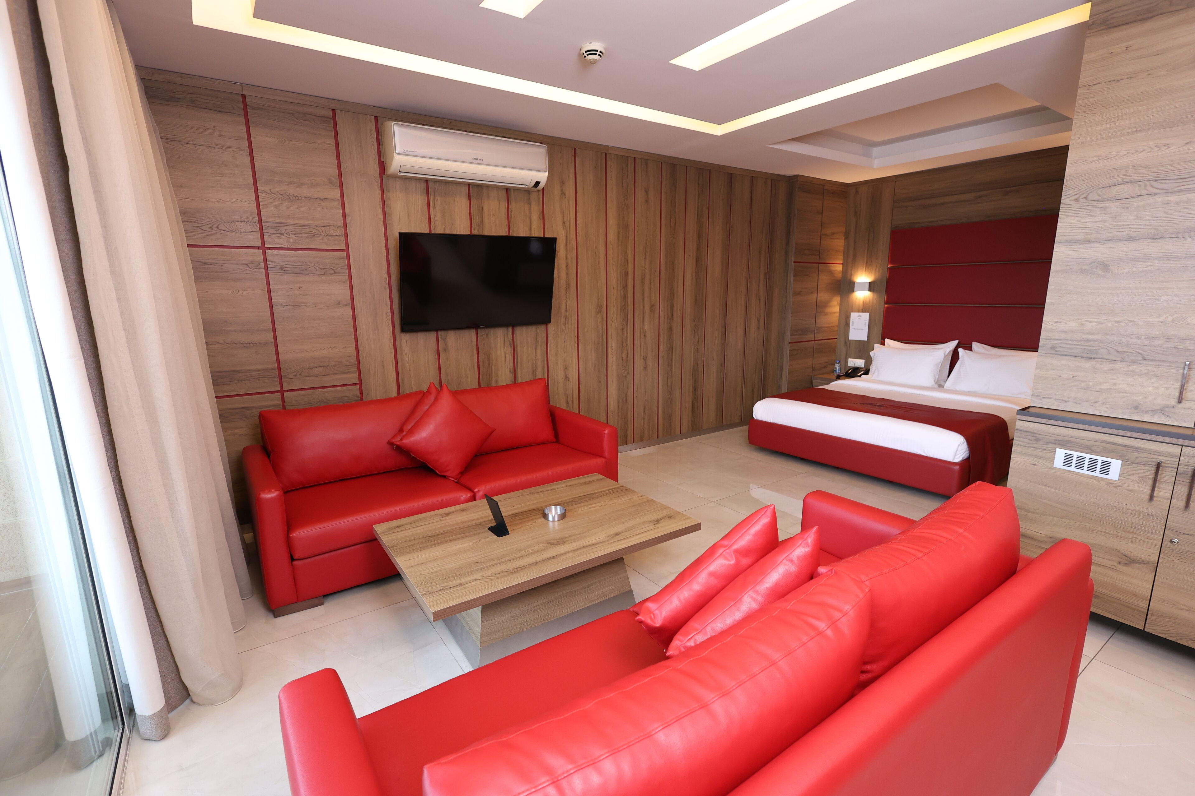 Una habitación de hotel moderna con un sofá rojo, una mesa de centro de madera, un televisor de pantalla plana y una cama con ropa blanca.