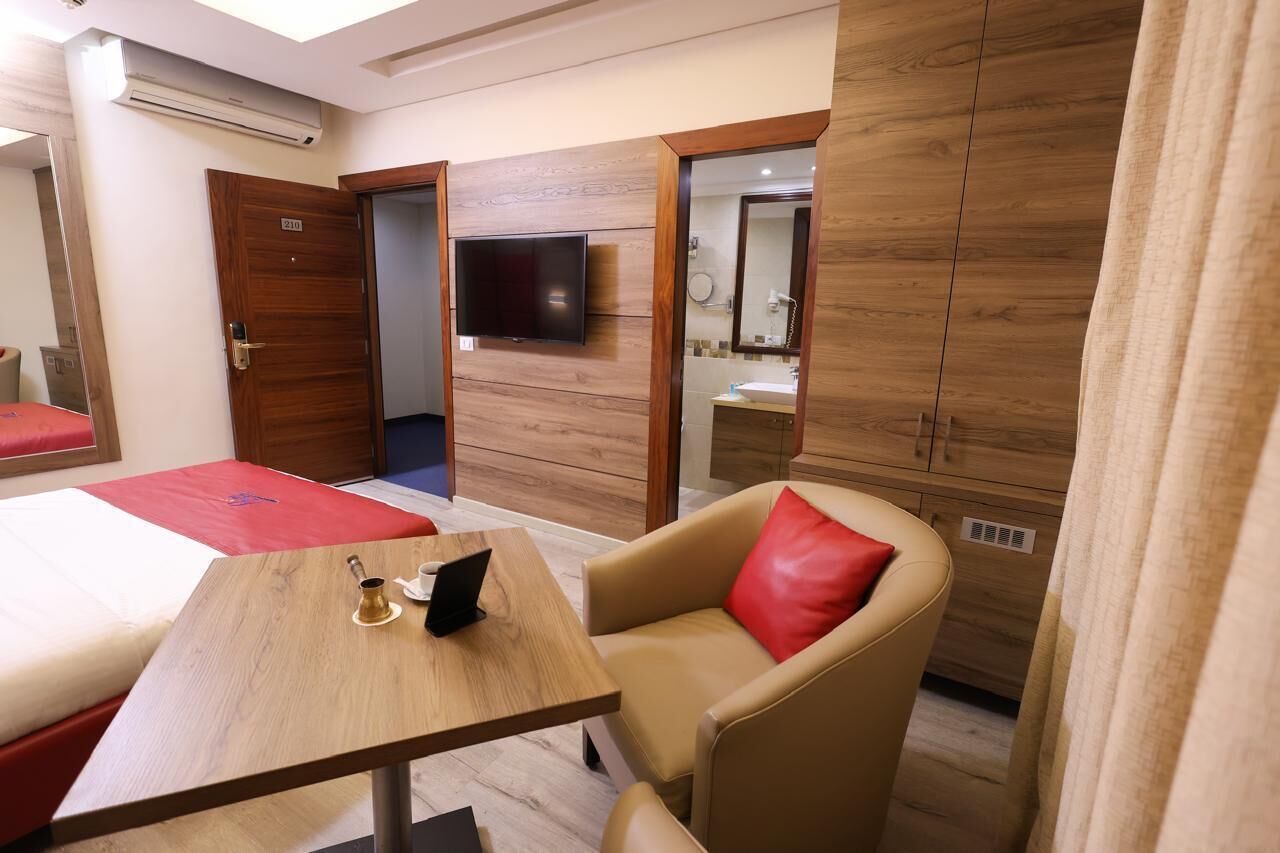 Une chambre d’hôtel avec un lit, une table en bois sur laquelle se trouve un ordinateur portable, une chaise avec un coussin rouge, un téléviseur fixé au mur et une salle de bain visible à travers une porte ouverte.