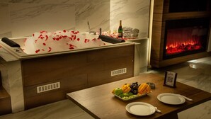 A bathroom with a marble sink, a bathtub with rose petals, a bottle of champagne, and a fruit platter on a wooden table.