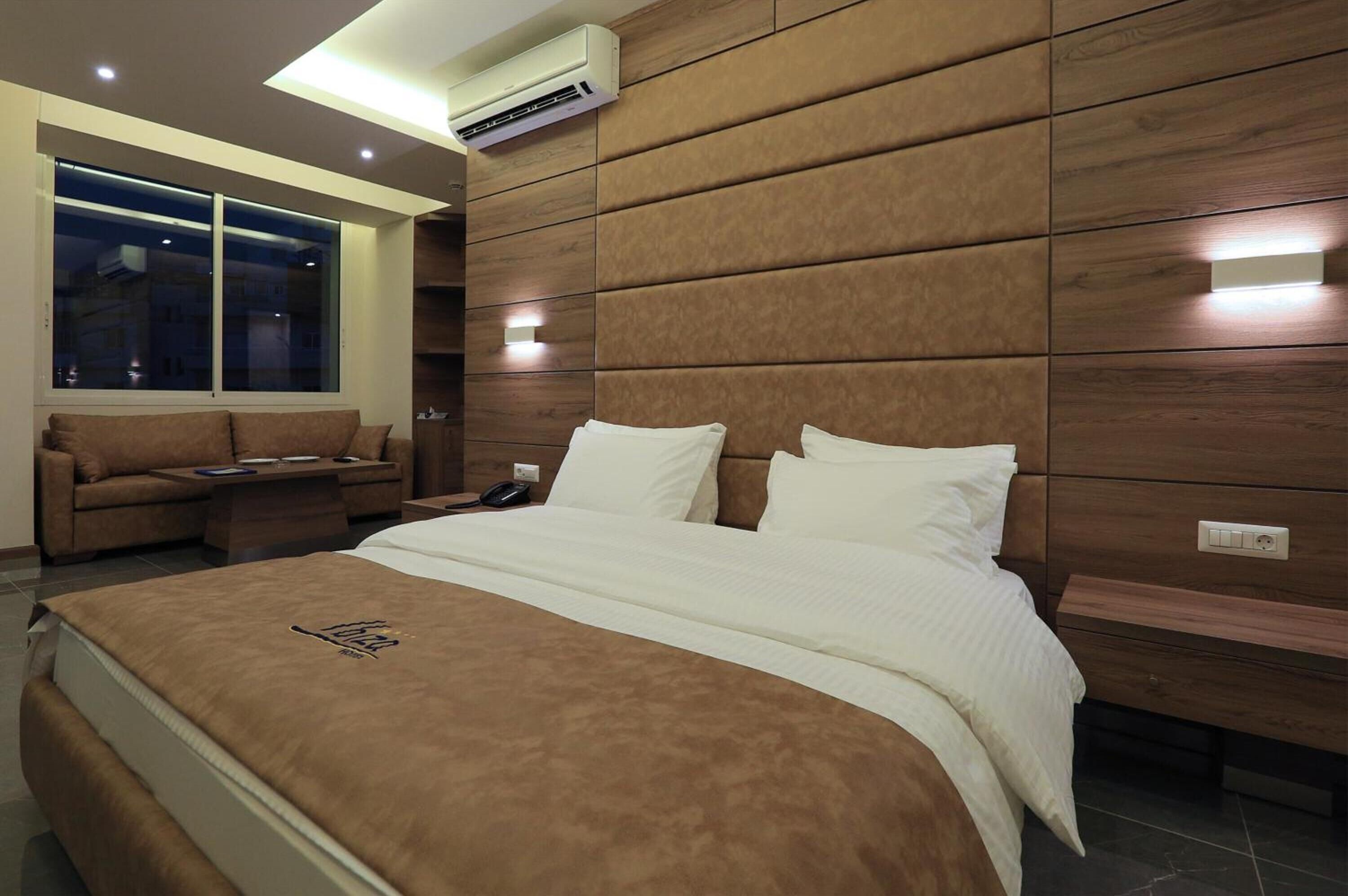 A modern hotel room with a large bed, a sofa, a nightstand, and a window with a view of the city at night.