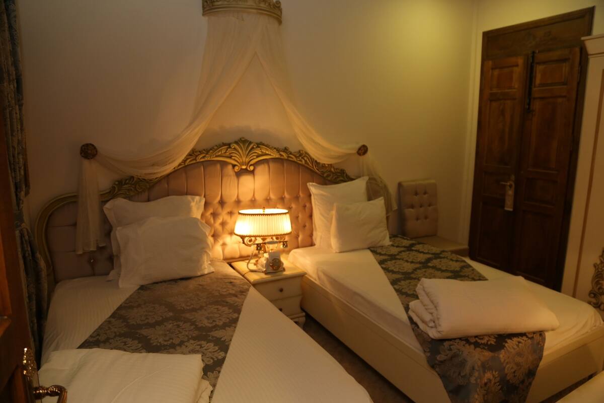 A double bed with a canopy, a bedside table with a lamp, and a wooden door in a hotel room.