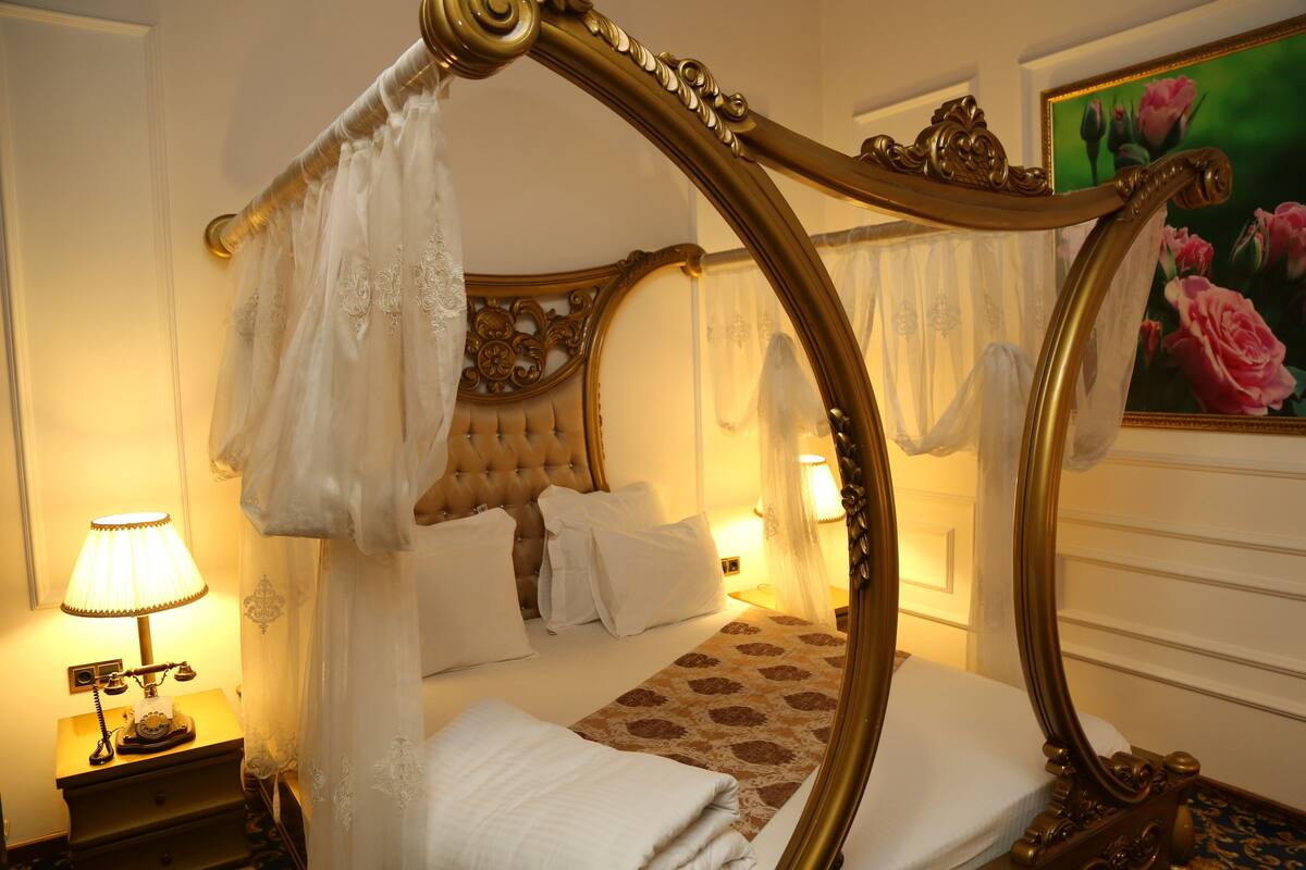 A four-poster bed with a canopy, a classic tufted headboard, and a floral-patterned bedspread. There is a bedside table with a lamp and a telephone.