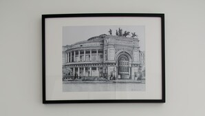 A framed black and white photograph of a classical building with columns and statues, mounted on a white wall above a glass-top table with two potted plants. - Appartamento Cavour 37 Palermo (Palermo)