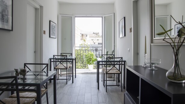 A modern dining area with a glass table and chairs, a potted plant, and a view of the city through an open door. - Appartamento Cavour 37 Palermo (Palermo)