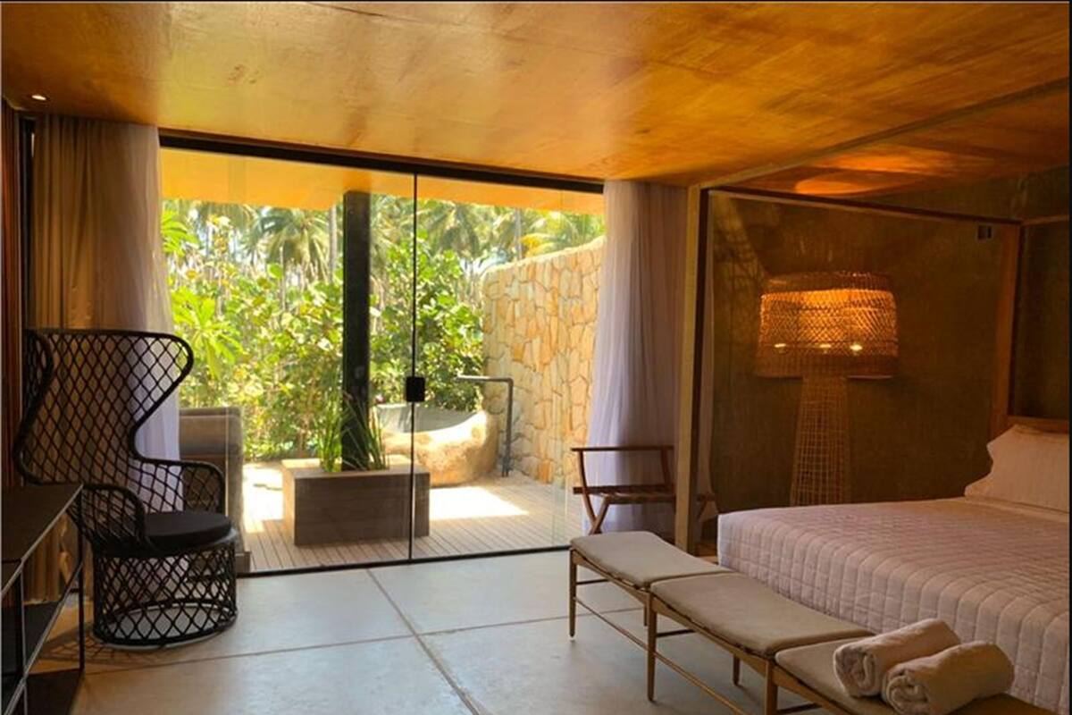 A modern bedroom with a large bed, a bench, a chair, and a view of greenery through a glass door.